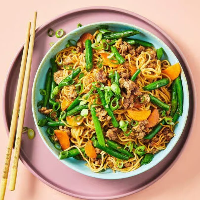 Noodles with ground meat, green beans, carrots, and scallions in a blue bowl with chopsticks.