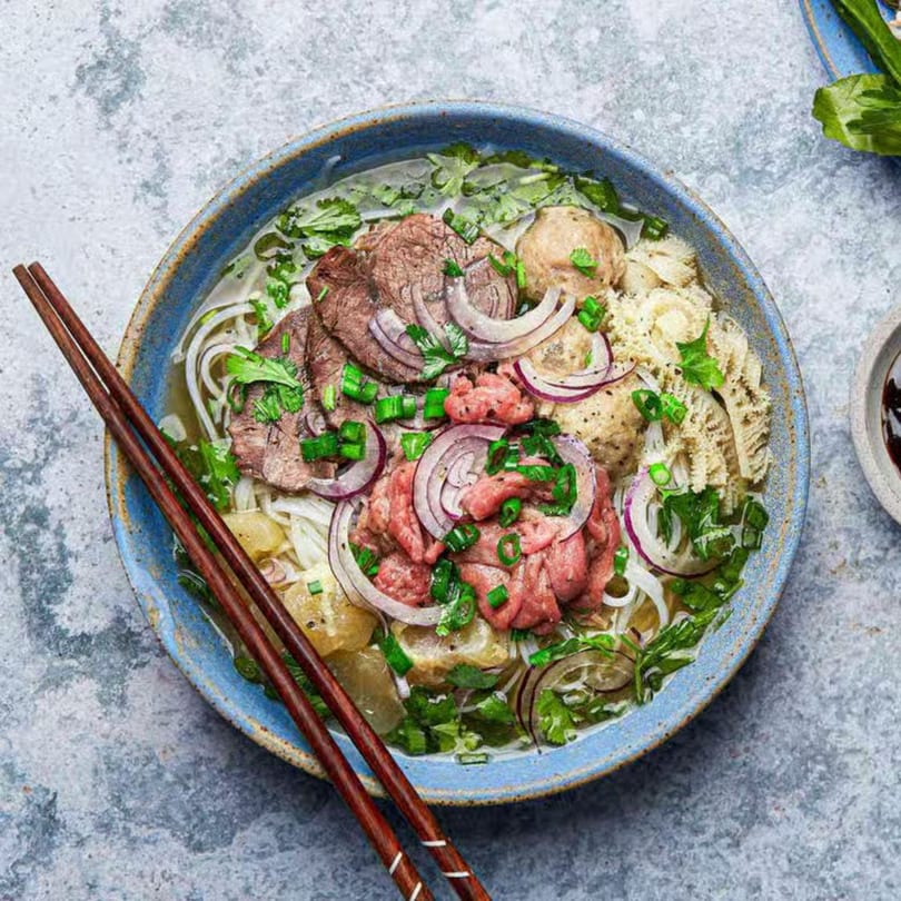 A blue bowl of Vietnamese pho with beef, noodles, onions, and herbs, with chopsticks.