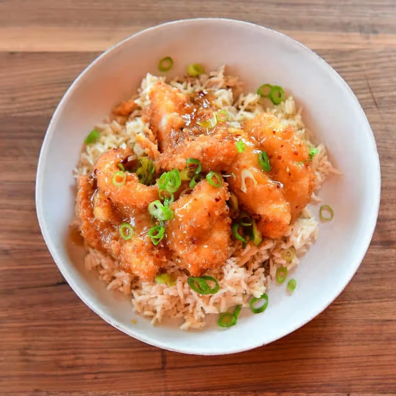 Crispy orange chicken and rice, garnished with green onions, in a white bowl on a wooden table.