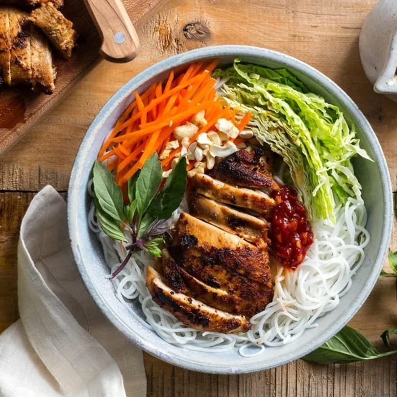 Grilled chicken, rice noodles, shredded carrots, lettuce, basil, and chili sauce in a bowl.